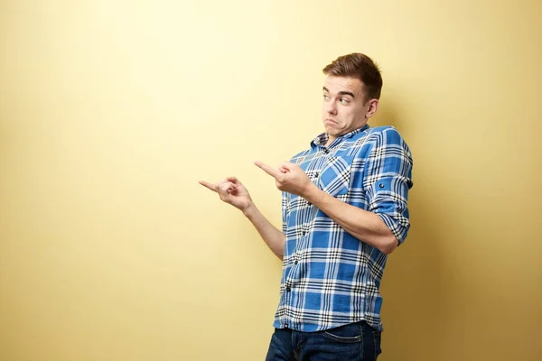 Förvånad killen klädd i en rutig skjorta och jeans står bredvid gul vägg i studion och punkter med händerna någonstans — Stockfoto