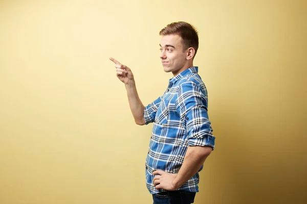 Cara surpreso vestido com uma camisa xadrez e jeans fica ao lado da parede amarela no estúdio — Fotografia de Stock