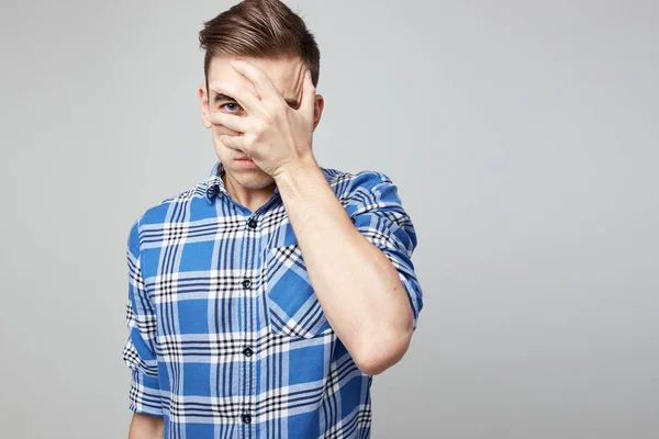 Rädd kille klädd i en rutig skjorta och jeans stänger ansiktet med handen på en vit bakgrund i studion — Stockfoto