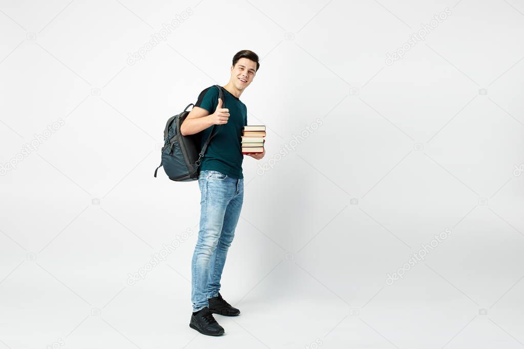 Funny dark-haired guy with a black backpack on his shoulder dressed in a dark t-shirt and jeans holds books in his hands on a white background