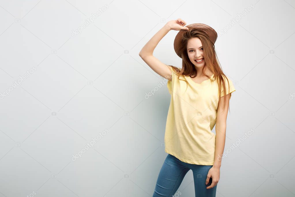 Smiling long haired girl dressed in a yellow t-shirt, jeans and hat stands on a white background in the studio
