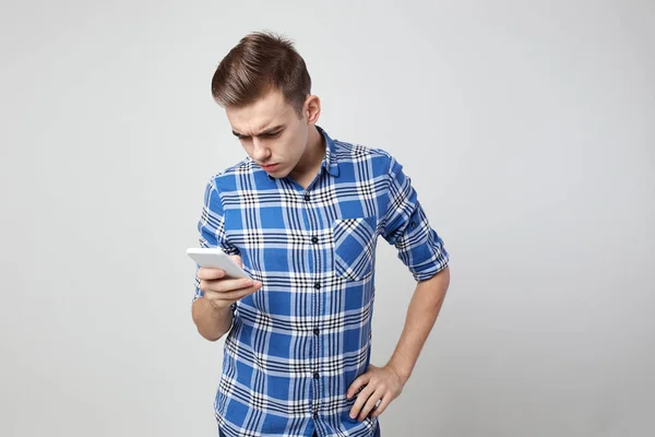 Cara surpreso vestido com uma camisa xadrez e jeans mantém o telefone celular em sua mão em um fundo branco no estúdio — Fotografia de Stock