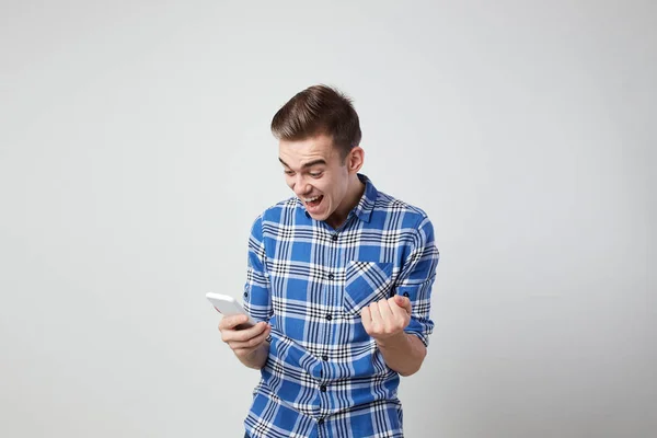 Extático cheio de energia cara vestido com uma camisa xadrez mantém o telefone celular em sua mão um fundo branco no estúdio — Fotografia de Stock
