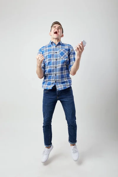 Extático cheio de energia cara vestido com uma camisa xadrez e jeans mantém o telefone celular em sua mão um fundo branco no estúdio e gritos alegremente — Fotografia de Stock