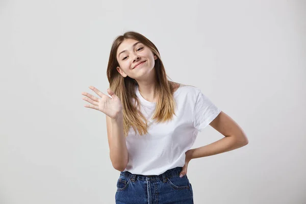 Chica amigable vestida con una camiseta blanca y jeans muestra hola en un fondo blanco en el estudio — Foto de Stock