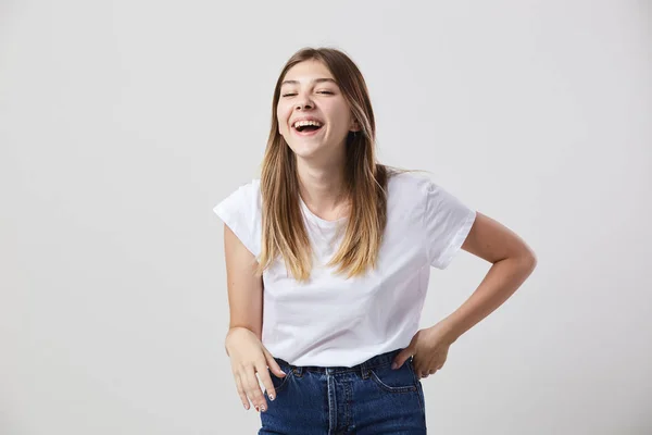 Дружня дівчина, одягнена в білу футболку і джинси, сміється на білому тлі в студії — стокове фото