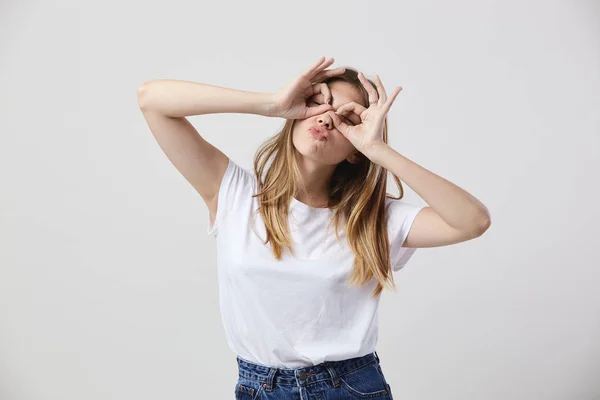 Grappig meisje gekleed in een wit t-shirt en jeans maakt bril met haar handen op haar ogen op een witte achtergrond in de studio — Stockfoto