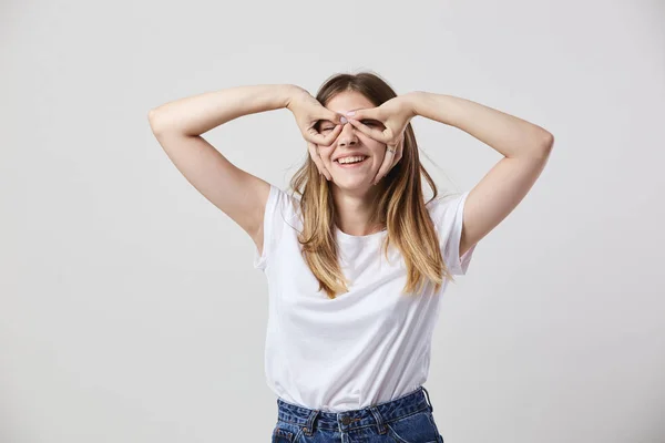 Lustiges Mädchen in weißem T-Shirt und Jeans macht Brille mit den Händen auf den Augen auf weißem Hintergrund im Studio — Stockfoto