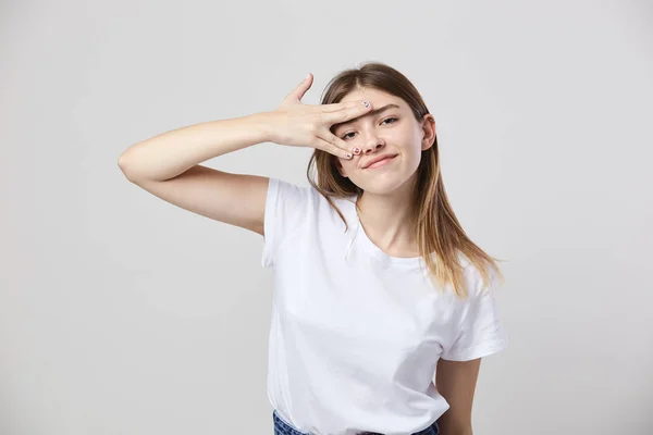 Vrolijk en rustig meisje gekleed in een wit t-shirt en jeans plaatst de vingers op het eerste gezicht op een witte achtergrond in de studio — Stockfoto