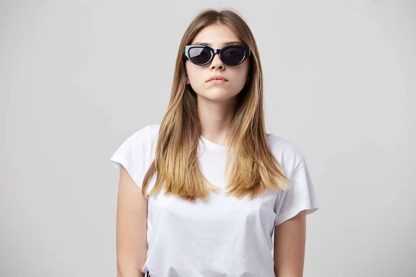 Серйозна дівчина в білій футболці і сонцезахисних окулярах на білому тлі в студії — стокове фото