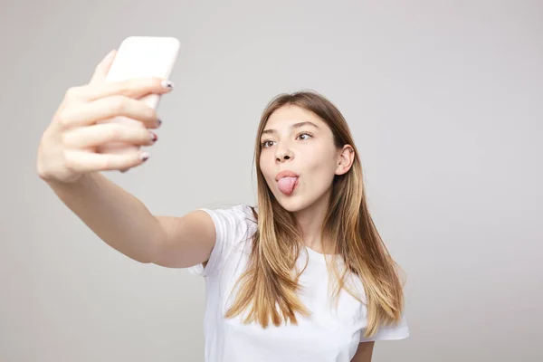 Щаслива дівчина в білій футболці показує, як язик робить селфі на білому тлі в студії — стокове фото