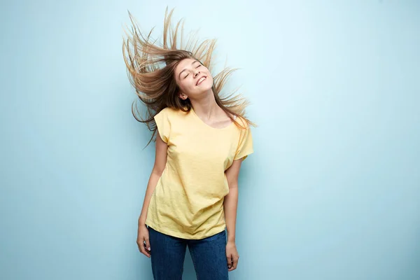 Menina sorridente com cabelo fluindo vestido com uma camiseta amarela e jeans está em um fundo azul no estúdio — Fotografia de Stock