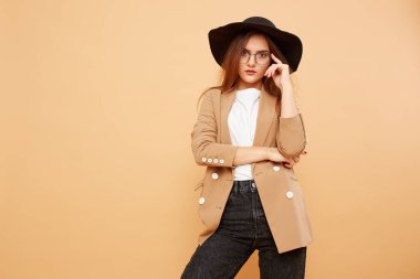 Beyaz t-shirt, kot pantolon ve bej ceketi giymiş başında siyah bir şapka ve gözlük uzun saçlı güzel esmer kız stüdyoda bej renkli arka plan üzerinde poz