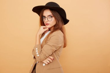 Şık beyaz t-shirt, kot pantolon ve bej ceketi giymiş başında siyah bir şapka ve gözlük uzun saçlı esmer kız stüdyoda bej renkli arka plan üzerinde poz