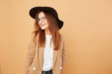 Gözlük ve başında siyah bir şapka uzun saçlı esmer kız gülümseyerek beyaz gömlek giymiş ve bej ceketi stüdyoda bej renkli arka plan üzerinde poz