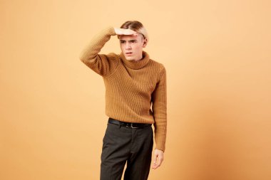 Genç sarışın adam hardal rengi kazak giymiş ve siyah pantolon bej renkli arka plan üzerinde stüdyoda alnındaki eliyle poz