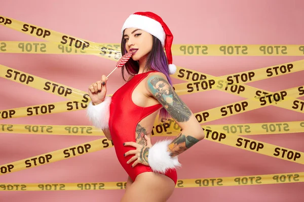 Meisje met tatoeages op haar lichaam gekleed in rood badpak, Santas hoed en witte vacht armbanden houden een lolly op de achtergrond van roze muur met de scotch-tapes met inscripties van woord "stop" — Stockfoto