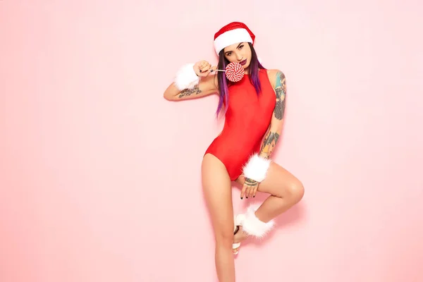 Girl with purple hair tips and tattoo on her arm dressed in red swimsuit, Santas hat and white fur bracelets holds a lollipop at her face on the background of pink wall