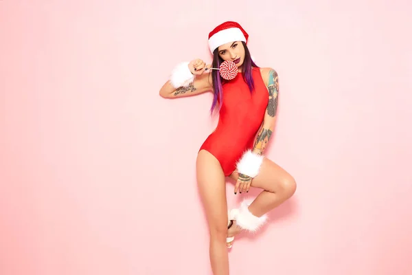 Ragazza con punte di capelli viola e tatuaggio sul braccio vestita in costume da bagno rosso, cappello di Babbo Natale e bracciali di pelliccia bianca tiene un lecca-lecca sul suo viso sullo sfondo della parete rosa — Foto Stock