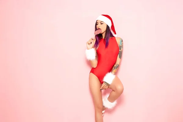 Girl with purple hair tips and tattoo on her arm dressed in red swimsuit, Santas hat and white fur bracelets licks a lollipop at her lips on the background of pink wall