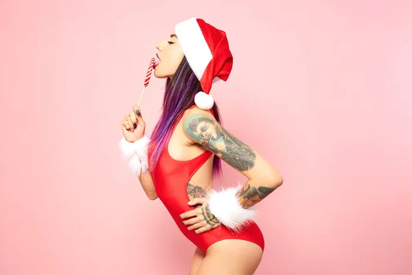 Girl with purple hair tips and tattoo on her arm dressed red swimsuit, Santas hat and white fur bracelets licks a lollipop in front of her face on the background of pink wall