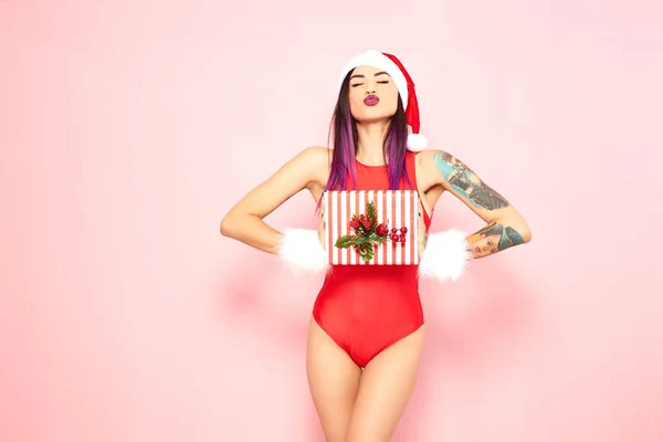 Girl with purple hair tips and tattoo on her arm dressed in red swimsuit, Santas hat and white fur bracelets holds a christmas gift in her hands on the background of pink wall .