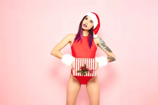 Girl with purple hair tips and tattoo on her arm dressed in red swimsuit, Santas hat and white fur bracelets holds a christmas gift in her hands on the background of pink wall