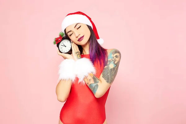 Girl with purple hair tips and tattoo on her arm dressed in red swimsuit, Santas hat and white fur bracelets holds a clock with five to twelve in hands on the background of pink wall