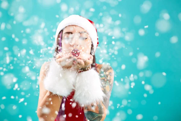 Heldere meisje met een tatoeage op haar arm gekleed in rood badpak, Santas hoed en witte vacht armbanden klappen sneeuwvlokken uit je handpalmen op de achtergrond van blauwe muur — Stockfoto