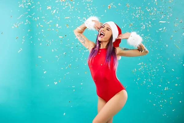 Genç kız kolunu dövme ile sesler konfeti ile mavi duvar arka plan üzerinde kırmızı mayo, Santas şapka ve beyaz kürk bilezik giyinmiş