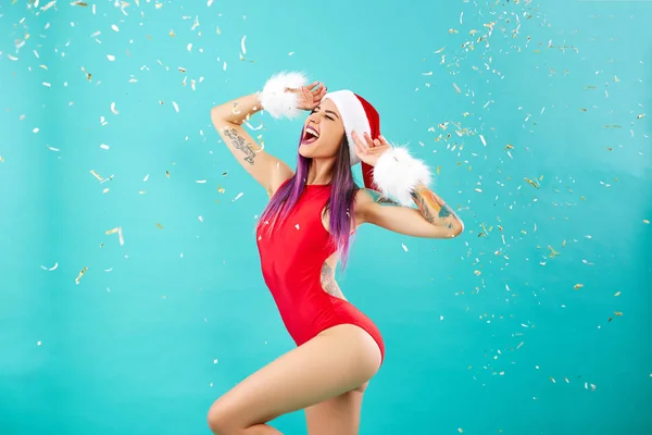 Jong meisje met een tatoeage op haar arm gekleed in rood badpak, Santas hoed en witte vacht armbanden geschreeuw op de achtergrond van blauwe muur met confetti — Stockfoto