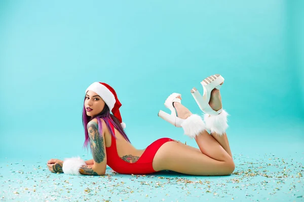 Charmantes Mädchen mit lila Haarspitzen und Tätowierungen auf den Armen in rotem Badeanzug, Weihnachtsmannmütze, weißen Fellarmbändern und High Heels liegt auf dem Boden, Konfetti auf blauem Hintergrund — Stockfoto