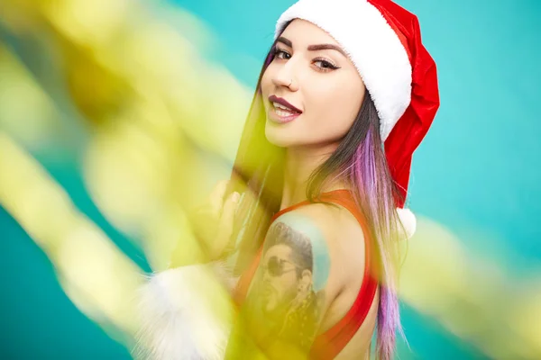 Charmante meisje met paars haar tips en tatoeage op haar arm gekleed in rood badpak, Santas hoed en witte vacht armbanden heeft plezier met confetti op de blauwe achtergrond — Stockfoto