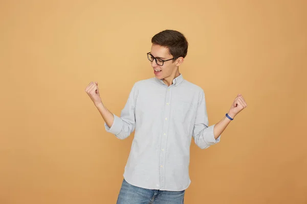 Young guy in glasses dressed in light blue shirt and jeans stands joyful on the beige background in the studio — Stock Photo, Image
