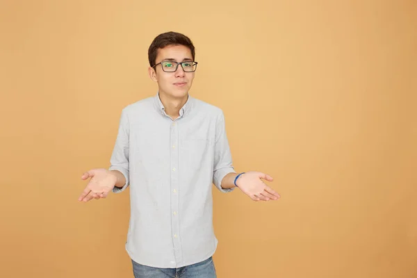 Jovem de óculos vestido de camisa azul clara e jeans fica com as mãos afastadas no fundo bege no estúdio — Fotografia de Stock