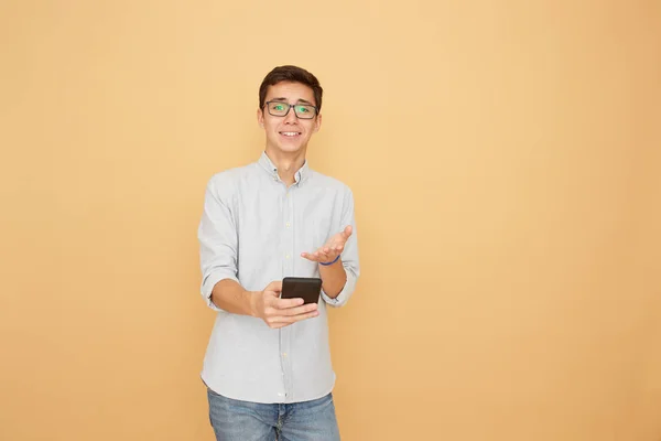 Jovem de óculos vestido com camisa azul clara e jeans segura um celular na mão no fundo bege no estúdio — Fotografia de Stock