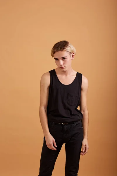 Young blond guy dressed in a black t-shirt and pants posing in the studio on the beige background