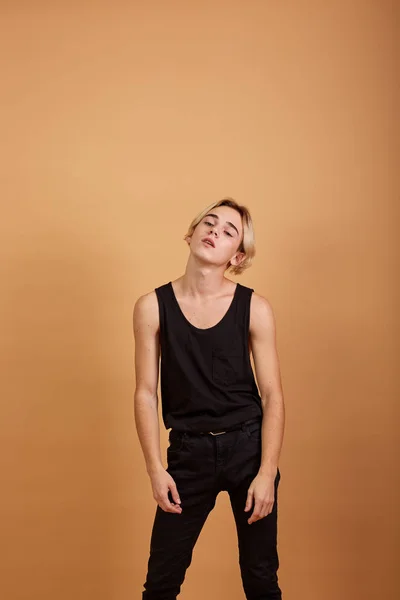 Young skinny blond guy dressed in a black tank top and pants posing in the studio on the beige background