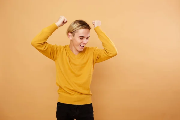 Joyful young blond guy dressed in yellow sweater is posing in the studio on the beige background