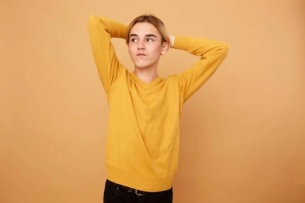Joven rubio vestido con suéter amarillo mantiene las manos en la cabeza sobre el fondo beige en el estudio —  Fotos de Stock