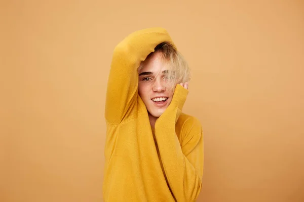 Blond guy dressed in yellow sweater is posing  with his hands on his face on the beige background in the studio