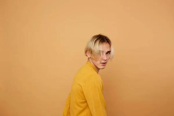 Stylish blond guy dressed in yellow sweater is posing on the beige background in the studio