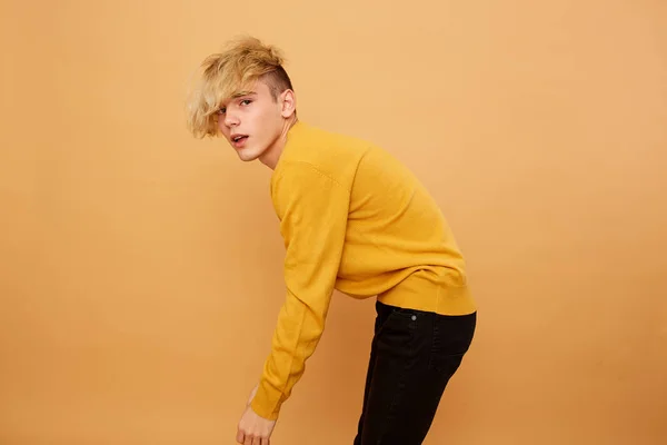Stylish blond guy dressed in yellow sweater and black jeans is posing on the beige background in the studio