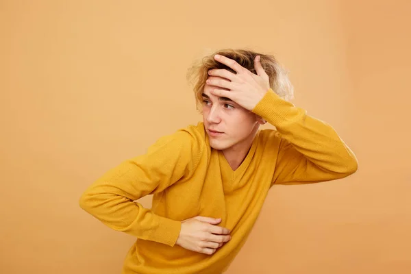 Stylish blond guy dressed in yellow sweater is posing with his hand on his head on the beige background in the studio