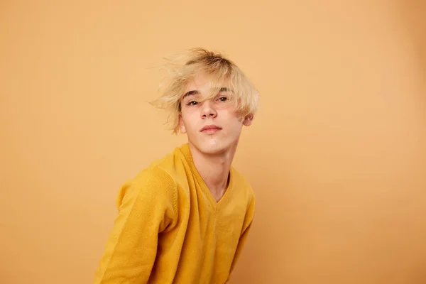 Young blond guy dressed in yellow sweater is posing on the beige background in the studio
