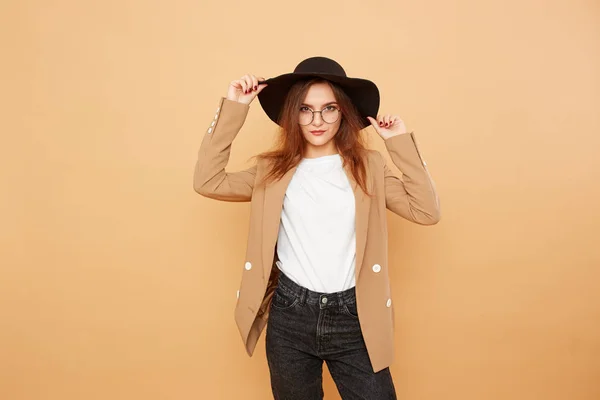 Pretty brunette girl with long hair in glasses and a black hat on her head dressed in white t-shirt, jeans and beige jacket is posing on the beige background in the studio — Stock Photo, Image
