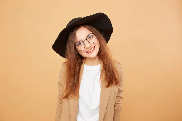 Усміхнена брюнетка дівчина з довгим волоссям в окулярах і чорний капелюх на голові в білій футболці і бежевий піджак позує на бежевому фоні в студії — стокове фото