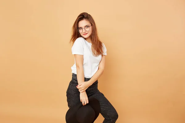 Brunette girl with long hair dressed in white t-shirt and black jeans is posing with black hat in her hand on the beige background in the studio — Stock Photo, Image