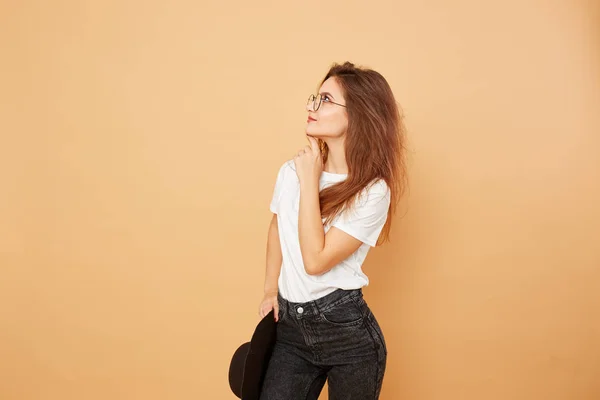 Chica morena con el pelo largo vestido con camiseta blanca y vaqueros negros está posando con sombrero negro en la mano sobre el fondo beige en el estudio — Foto de Stock