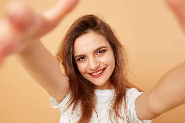 Brunette meisje met lang haar gekleed in witte t-shirt maakt een selfie op de beige achtergrond in de studio — Stockfoto